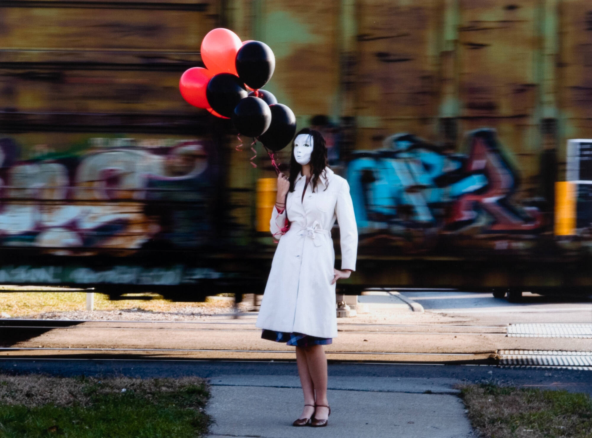 Color photograph of a woman wearing a white coat, a white mask covering her face, holding black and red balloons.